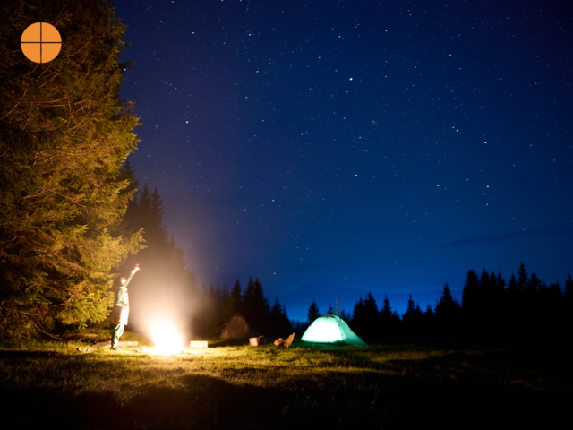 Person poiting at the night sky in the middle of the forrest, a fire and camping tent can also been seen in the photo. 