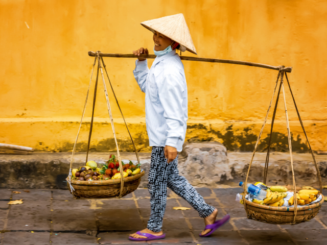 Traditionaly dresses vietnamese street vendor woman carrying her goods in two baskets hanging at each end of a bamboo pole.